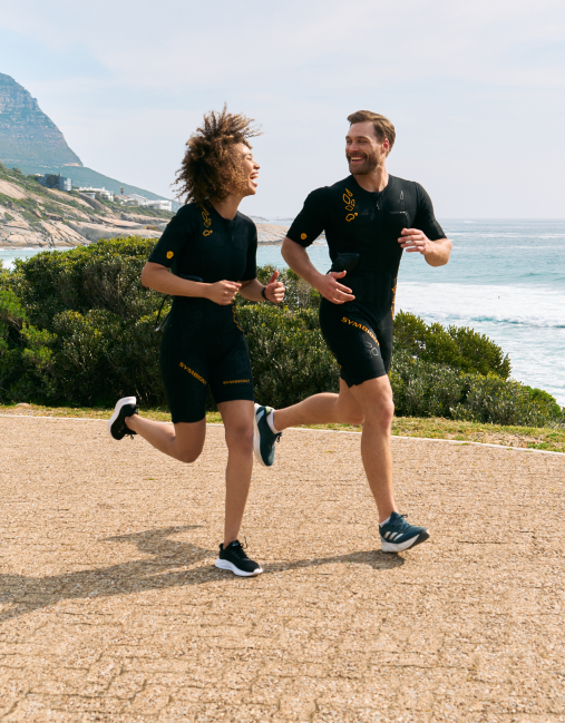 Two people in EMS suits running on the beach, smiling and enjoying the freedom and flexibility of their EMS training experience.