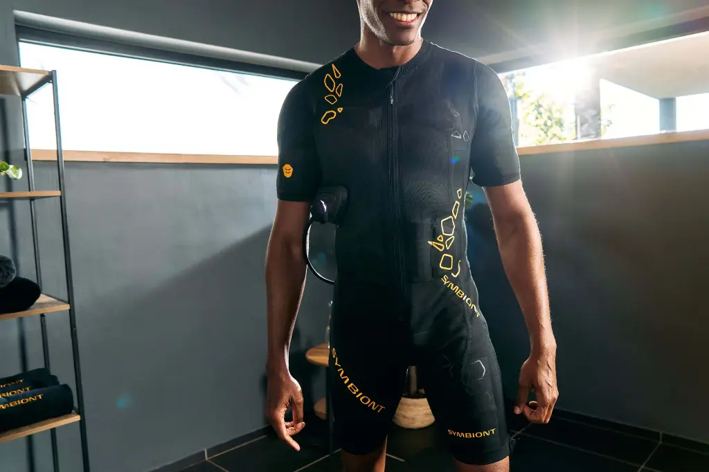 Premium EMS Equipment for Your Success – Full-Detail View of SYMBIONT EMS Suit Person standing in the EMS suit, enjoying the positive feeling of the EMS training experience. Focus on a full view of the suit's premium details and advanced features.
